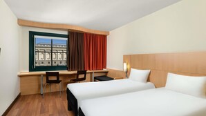 Quarto de hotel com duas camas, uma escrivaninha, duas cadeiras, uma janela com cortinas e vista para um edifício.
