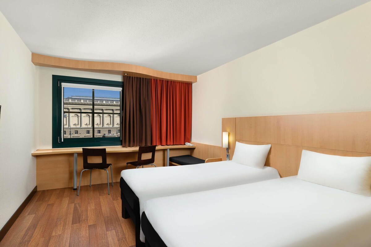 A hotel room with two beds, a desk, two chairs, a window with curtains, and a view of a building.