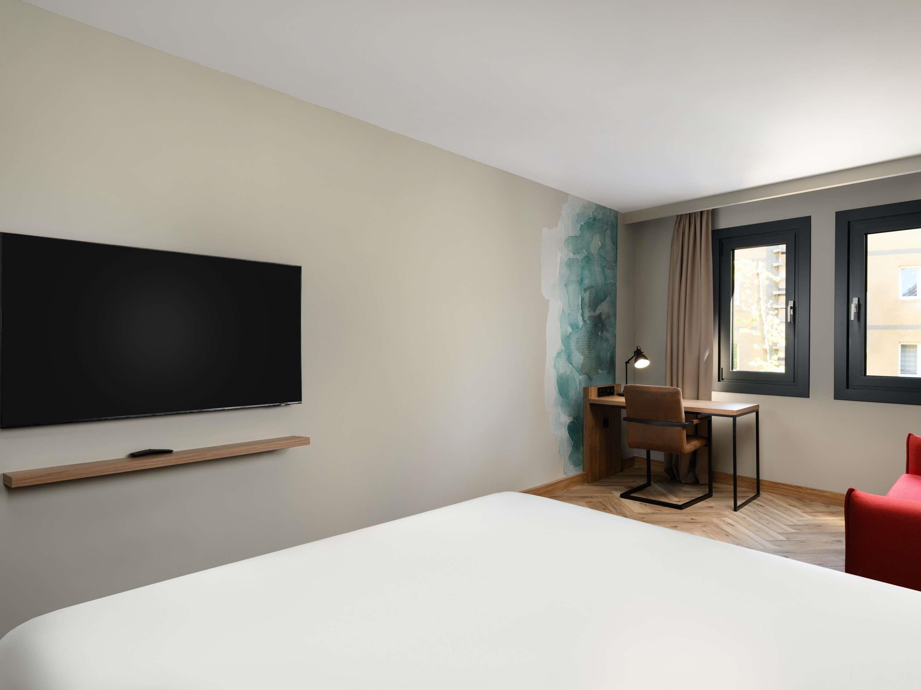 A hotel room with a bed, a desk, a chair, a TV, and windows.