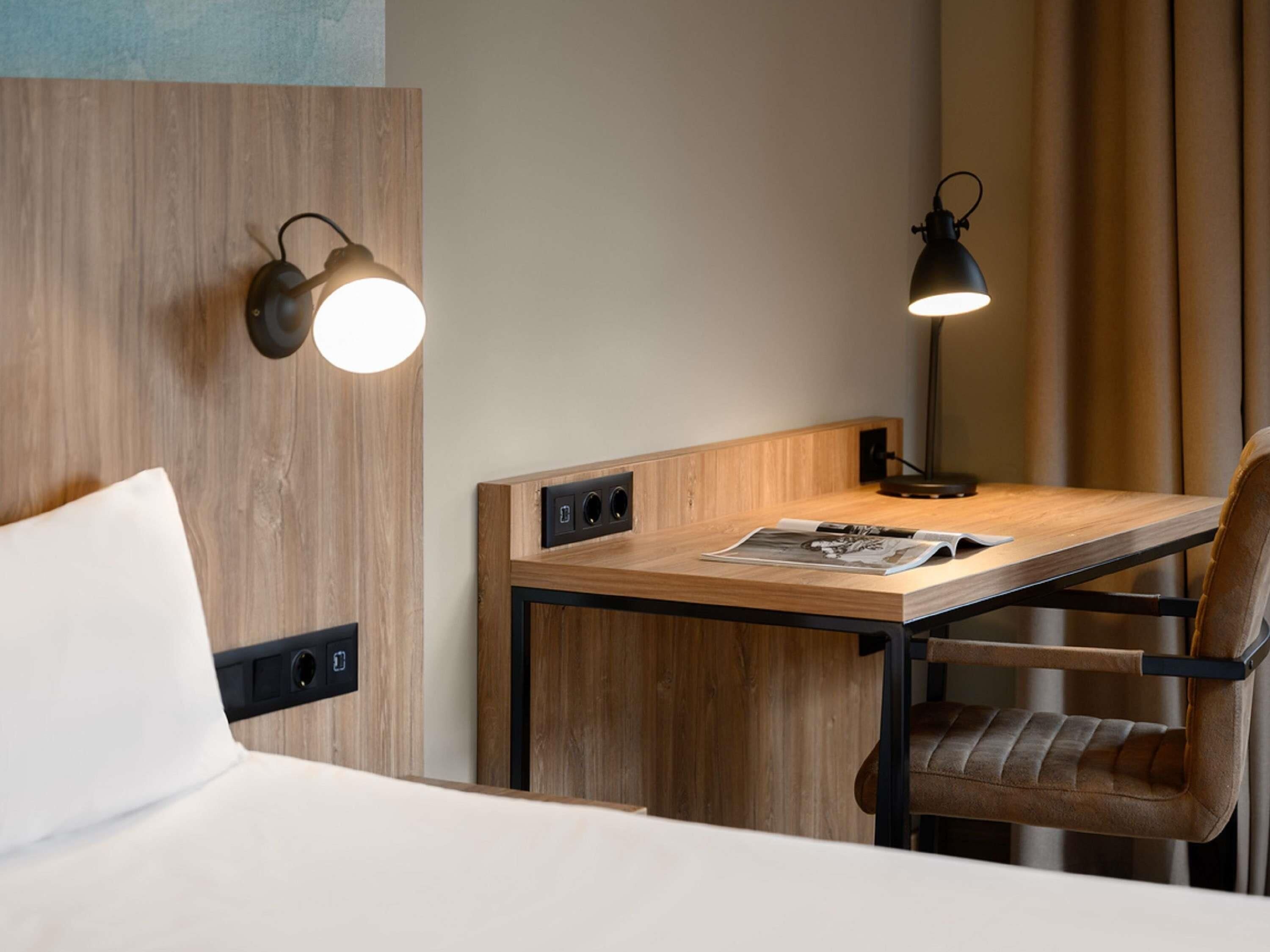 A hotel room with a bed, a desk with a chair, two wall-mounted lamps, and a window with curtains.