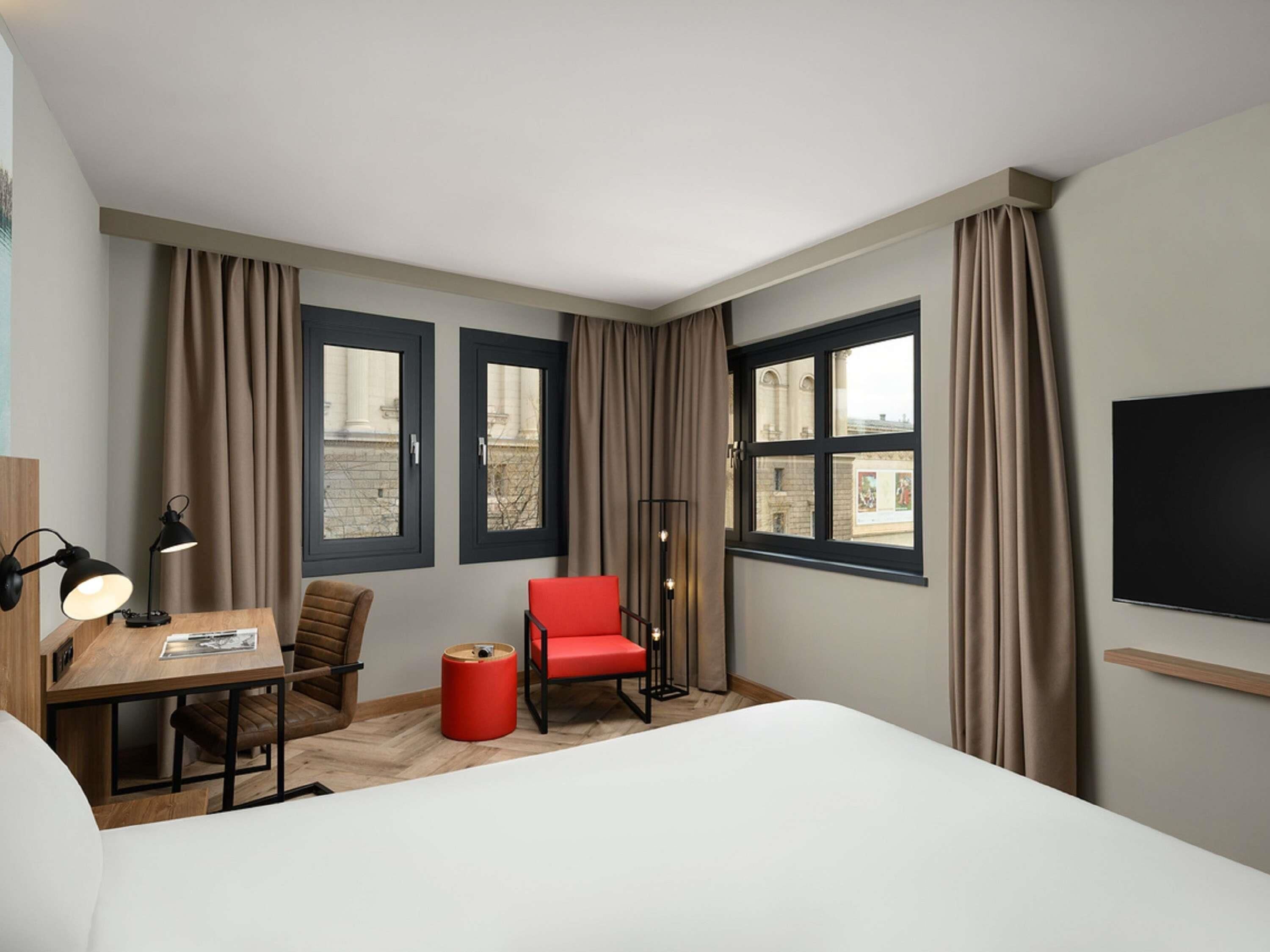 Habitación de hotel con cama, escritorio con lámpara, una silla roja y dos ventanas con cortinas.