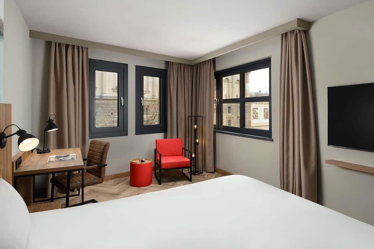 A hotel room with a bed, a desk with a lamp, a red chair, and two windows with curtains.