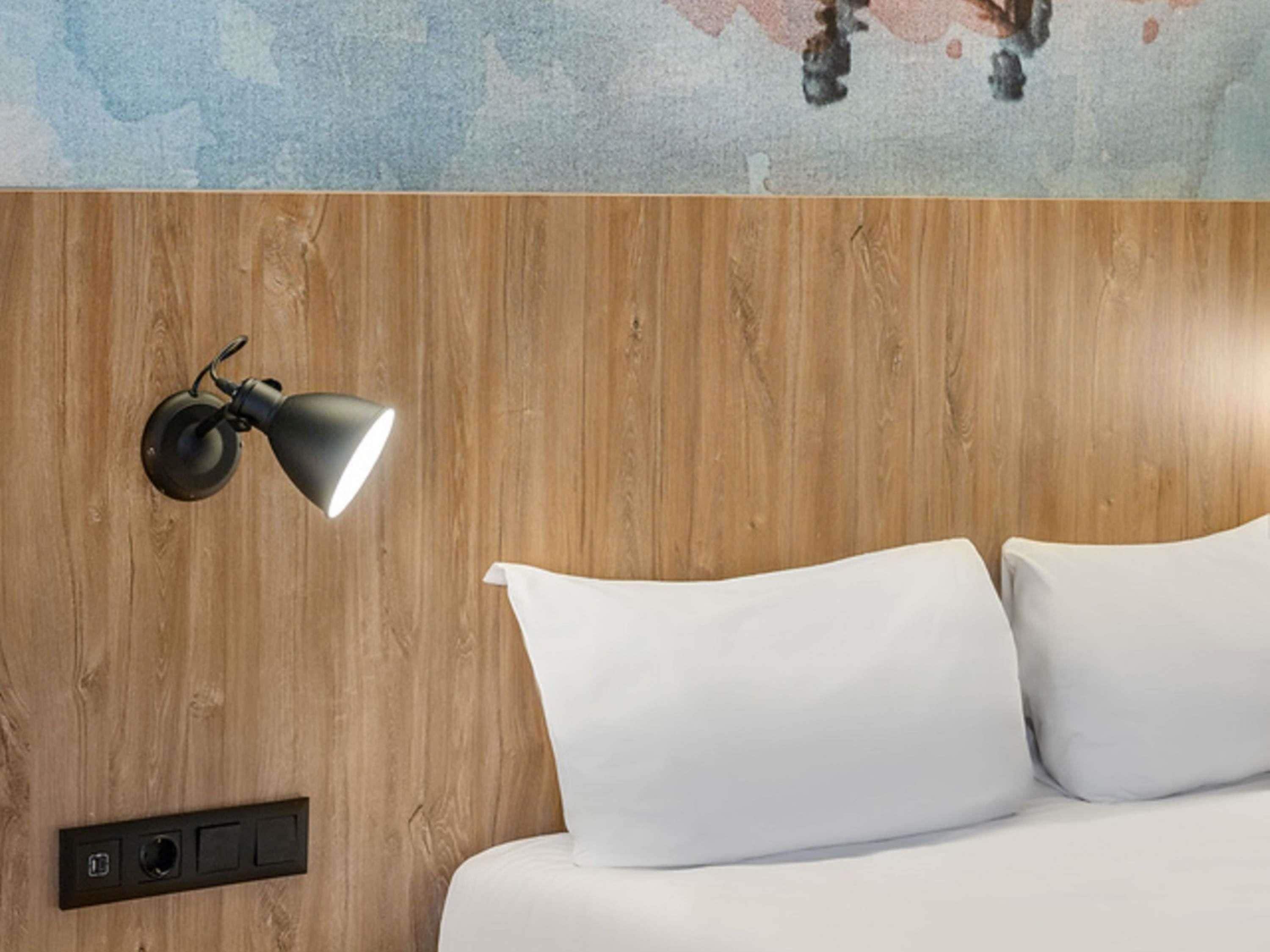A bed with white linens, a wooden headboard, a wall-mounted lamp, and a control panel.