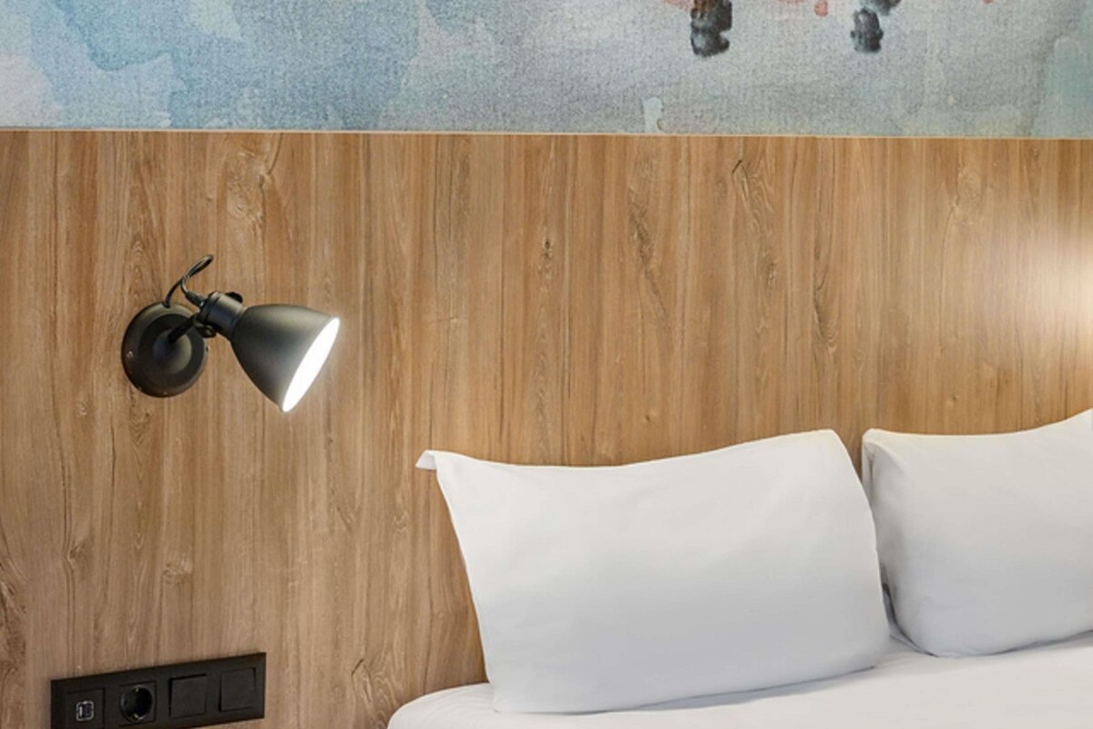 Ein Bett mit weißen Leinen, einem Holz-Kopfteil, einer Wandlampe und einem Bedienfeld.