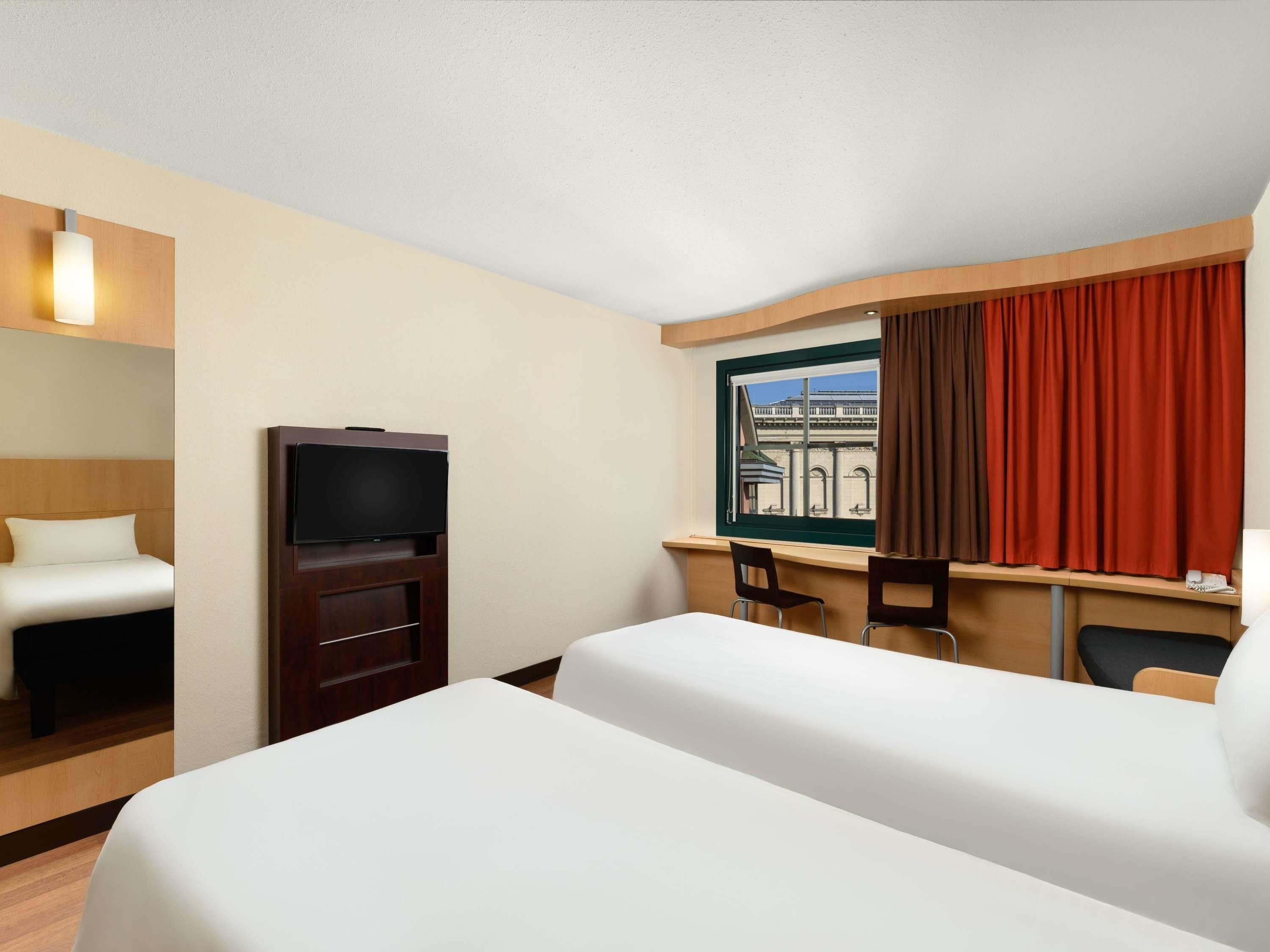 Ein Hotelzimmer mit einem Bett, einem Fernseher, einem Schreibtisch mit zwei Stühlen und einem Fenster mit Vorhängen.