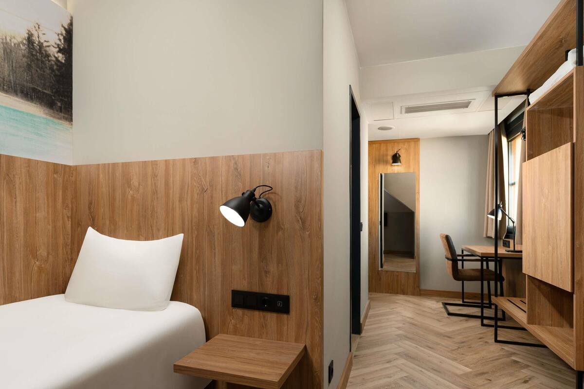 Moderni hotellihuone, jossa on puinen pääty, valkoisilla tekstiileillä varustettu sänky, yöpöytä, työpöytä tuolilla ja peili.