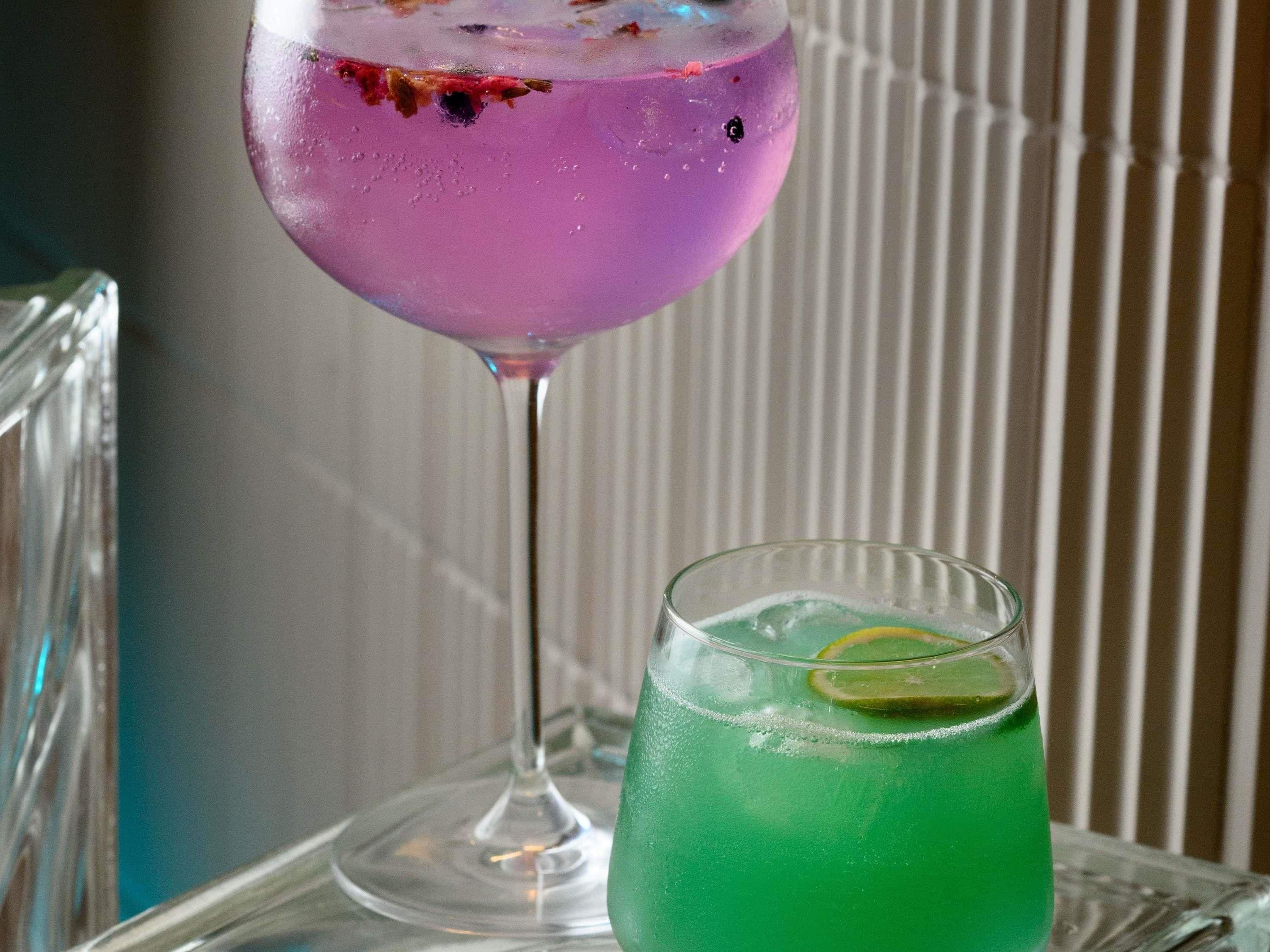 A purple cocktail in a stemmed glass and a green drink in a short glass with a lime slice, both on a clear glass table.
