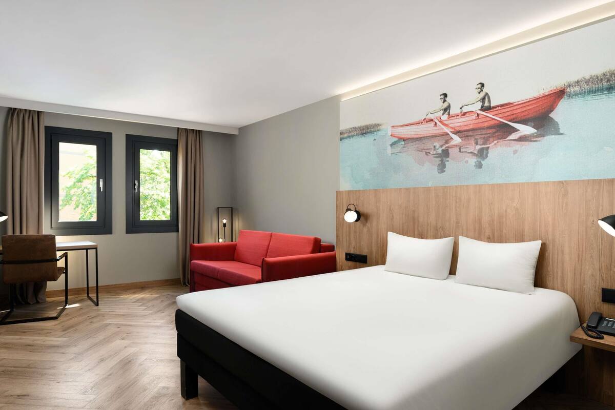 Hotellihuone, jossa on suuri sänky, punainen nojatuoli, työpöytä ja seinällä oleva maalaus, jossa on kuvattuna vene.