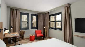 Habitación de hotel con cama, escritorio con lámpara, una silla roja y dos ventanas con cortinas.