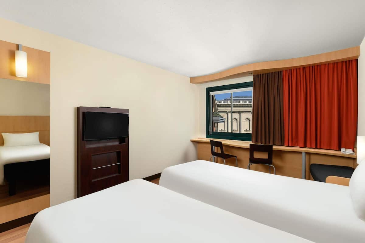 A hotel room with a bed, a TV, a desk with two chairs, and a window with curtains.
