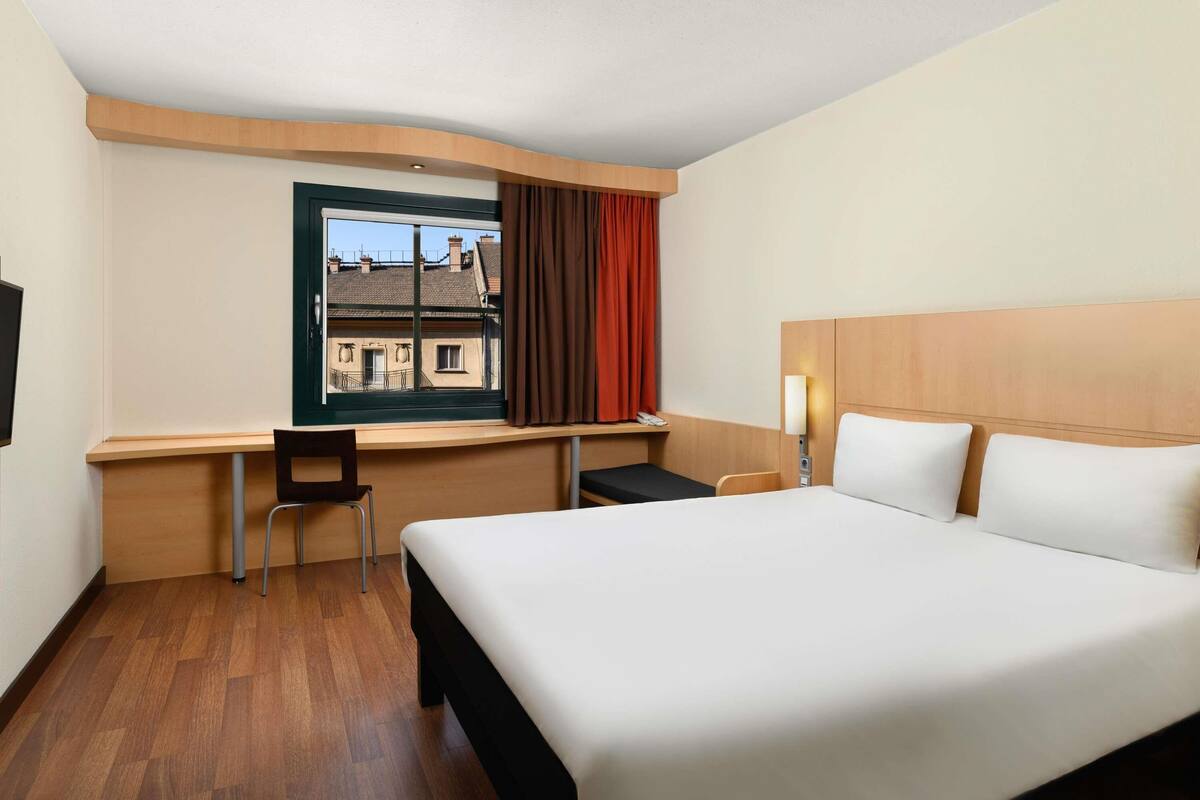 Hotellihuone, jossa on sänky, työpöytä, tuoli, ikkuna verhoilla ja näkymä rakennuksiin.