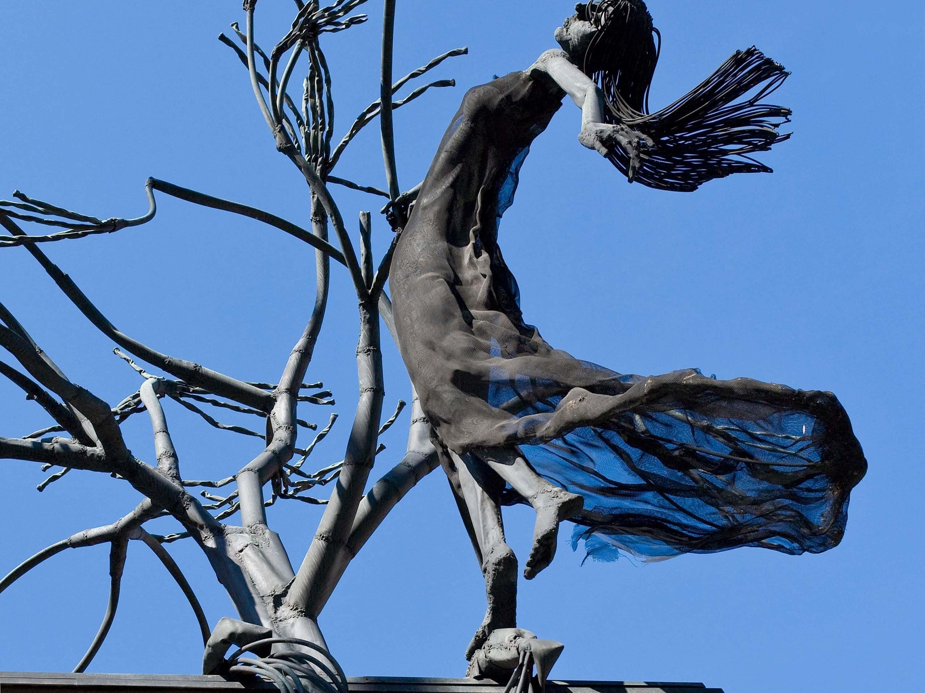 Une sculpture représentant une figure aux bras tendus, vêtue d’un tissu flottant, se détachant sur un ciel bleu clair.