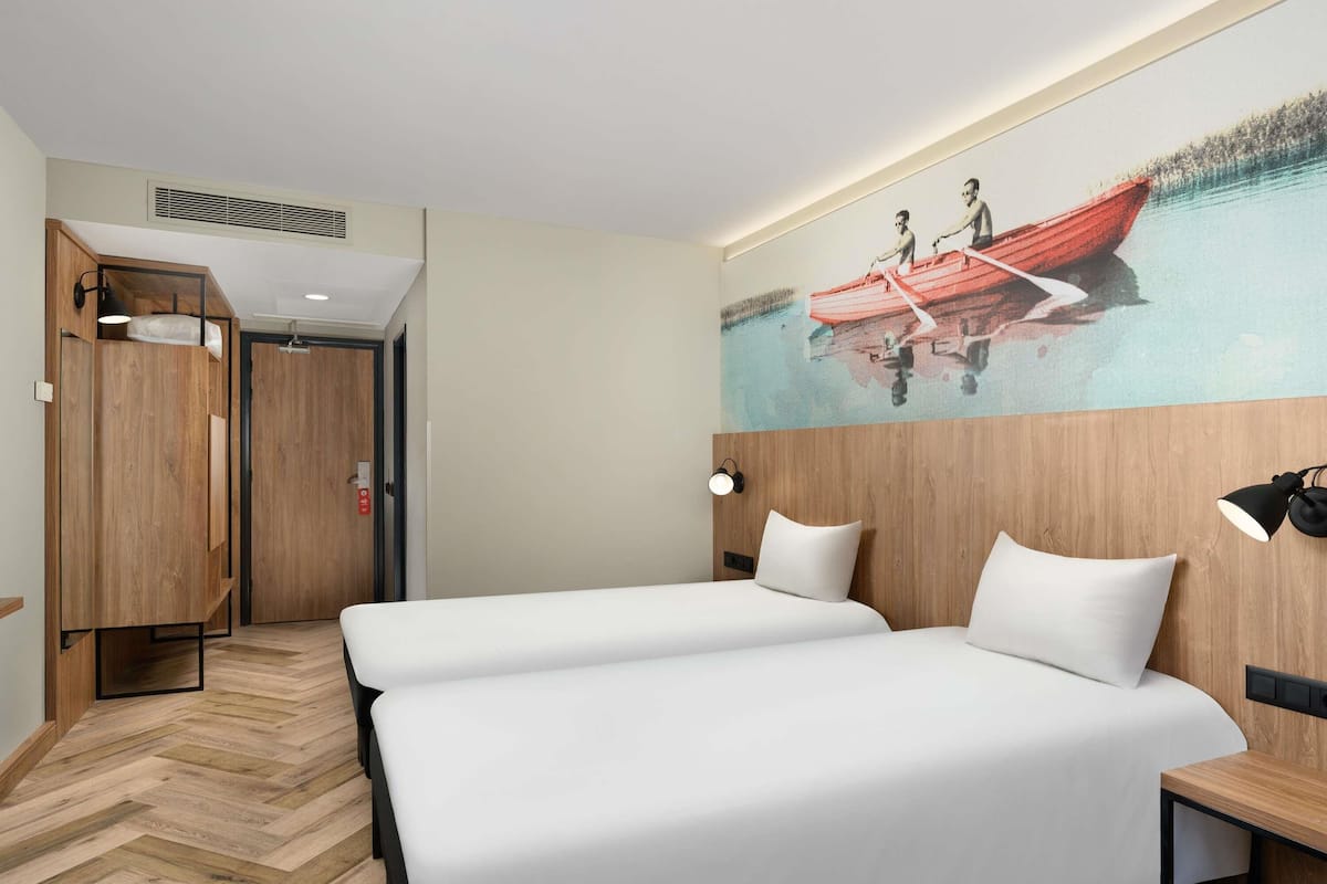 A hotel room with two beds, a wooden headboard, a wall-mounted lamp, and a painting of a red boat.