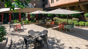 An outdoor patio area with tables and chairs, surrounded by greenery and a building with multiple windows. - ibis Styles Budapest Heroes Square (Budapest)