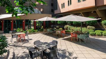 Un patio al aire libre con mesas y sillas, rodeado de follaje y un edificio con múltiples ventanas.