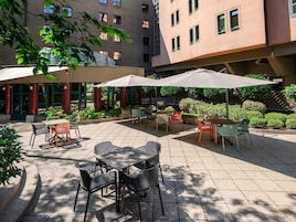 Un patio al aire libre con mesas y sillas, rodeado de follaje y un edificio con múltiples ventanas.