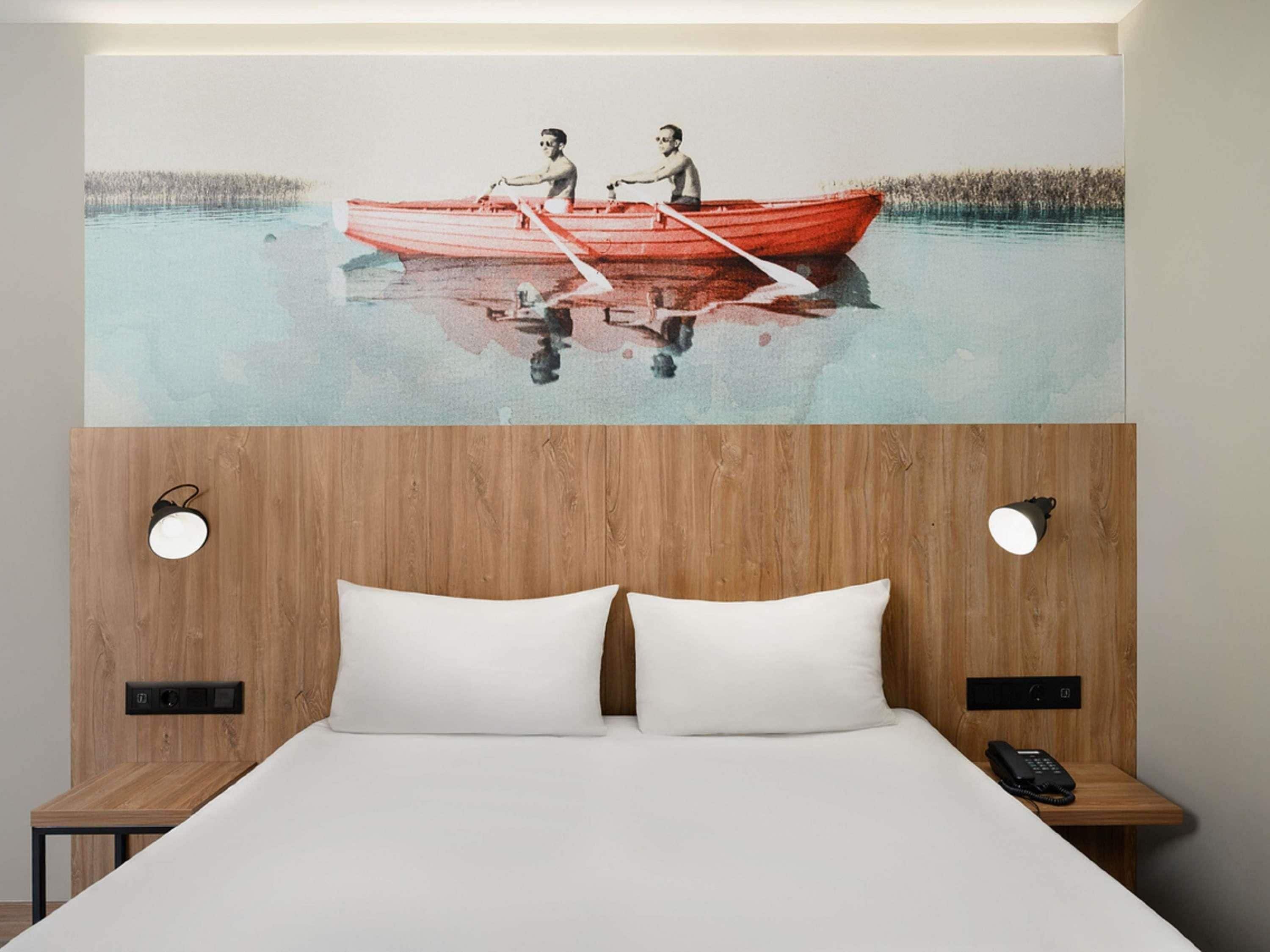 A hotel room with a wooden headboard, two white pillows, a wooden bedside table, and a painting of two people in a red boat on a calm water body.