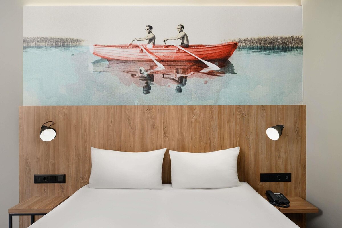 Ein Hotelzimmer mit einem Holzkopfteil, zwei weißen Kissen, einem Nachttisch aus Holz und einem Gemälde von zwei Personen in einem roten Boot auf ruhigem Wasser.