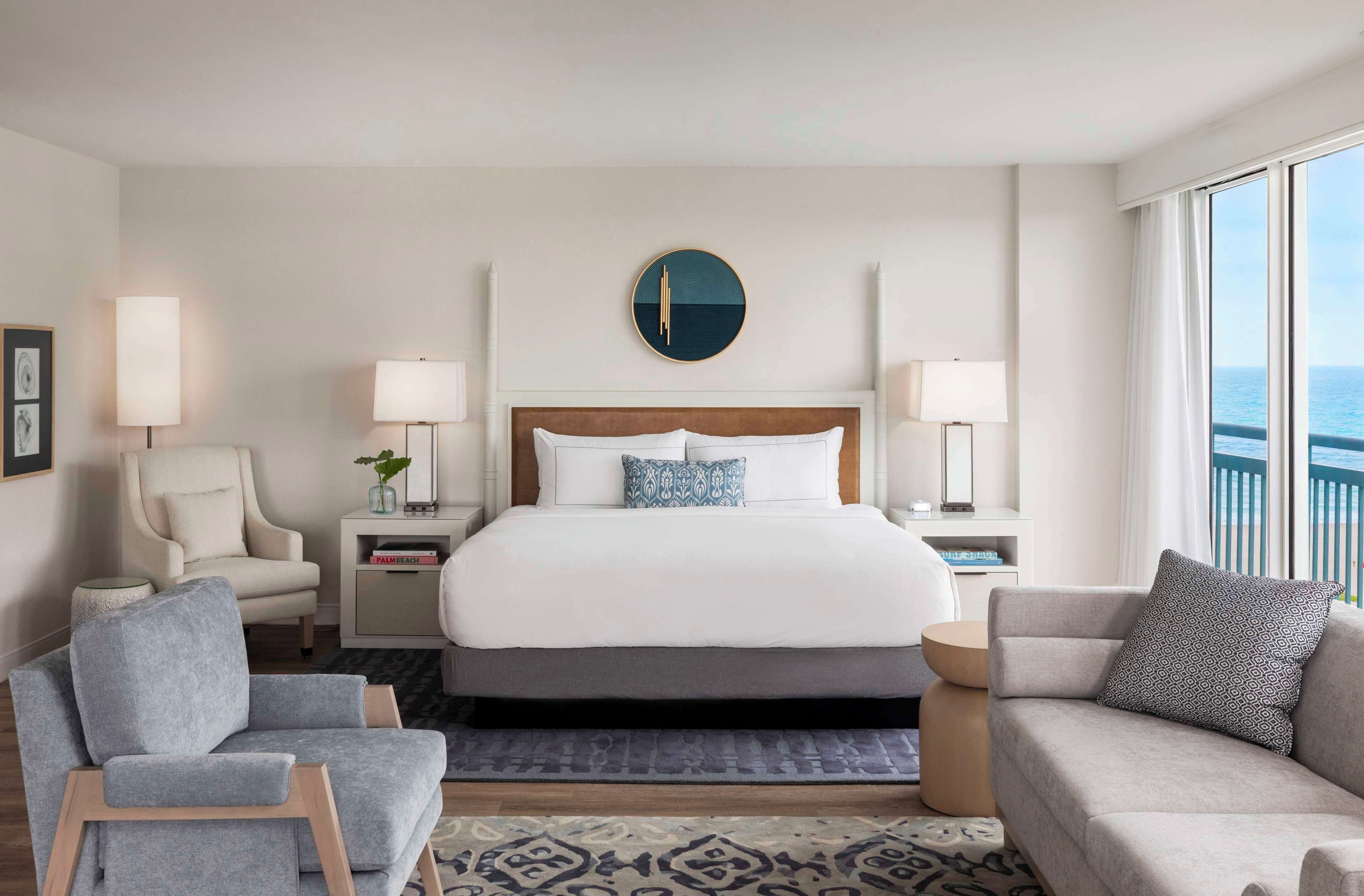 A hotel room with a large bed, two bedside tables with lamps, a sofa, a chair, and a view of the sea.