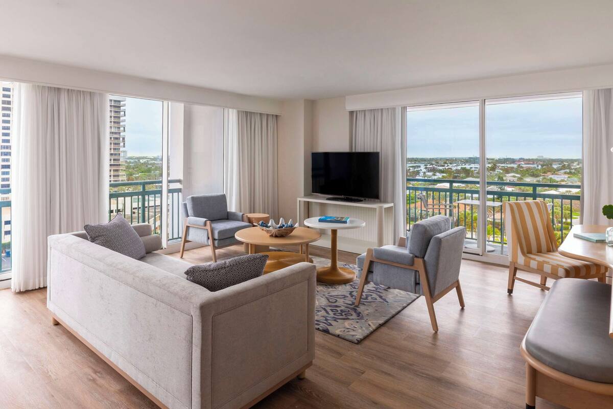 A modern living room with a sofa, chairs, a coffee table, a TV, and a balcony with a view.