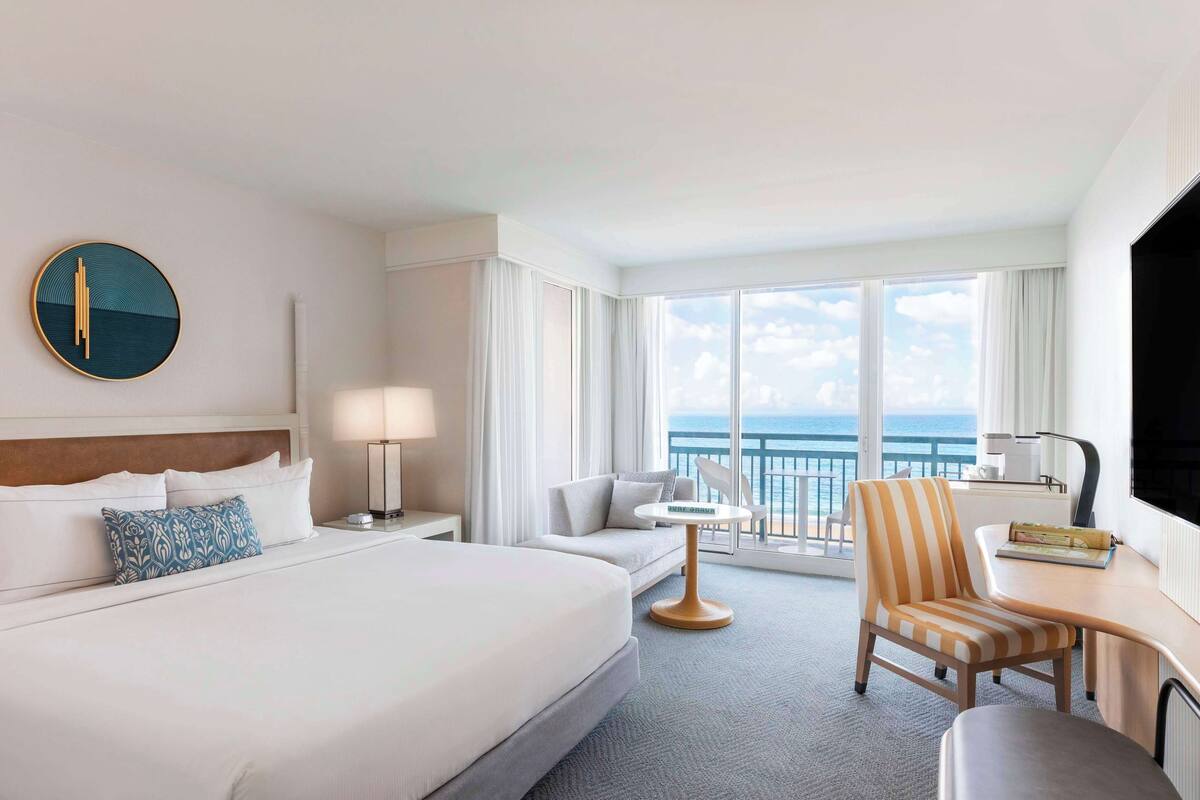 A hotel room with a large bed, a desk, a chair, a small table, and a view of the ocean through large windows.