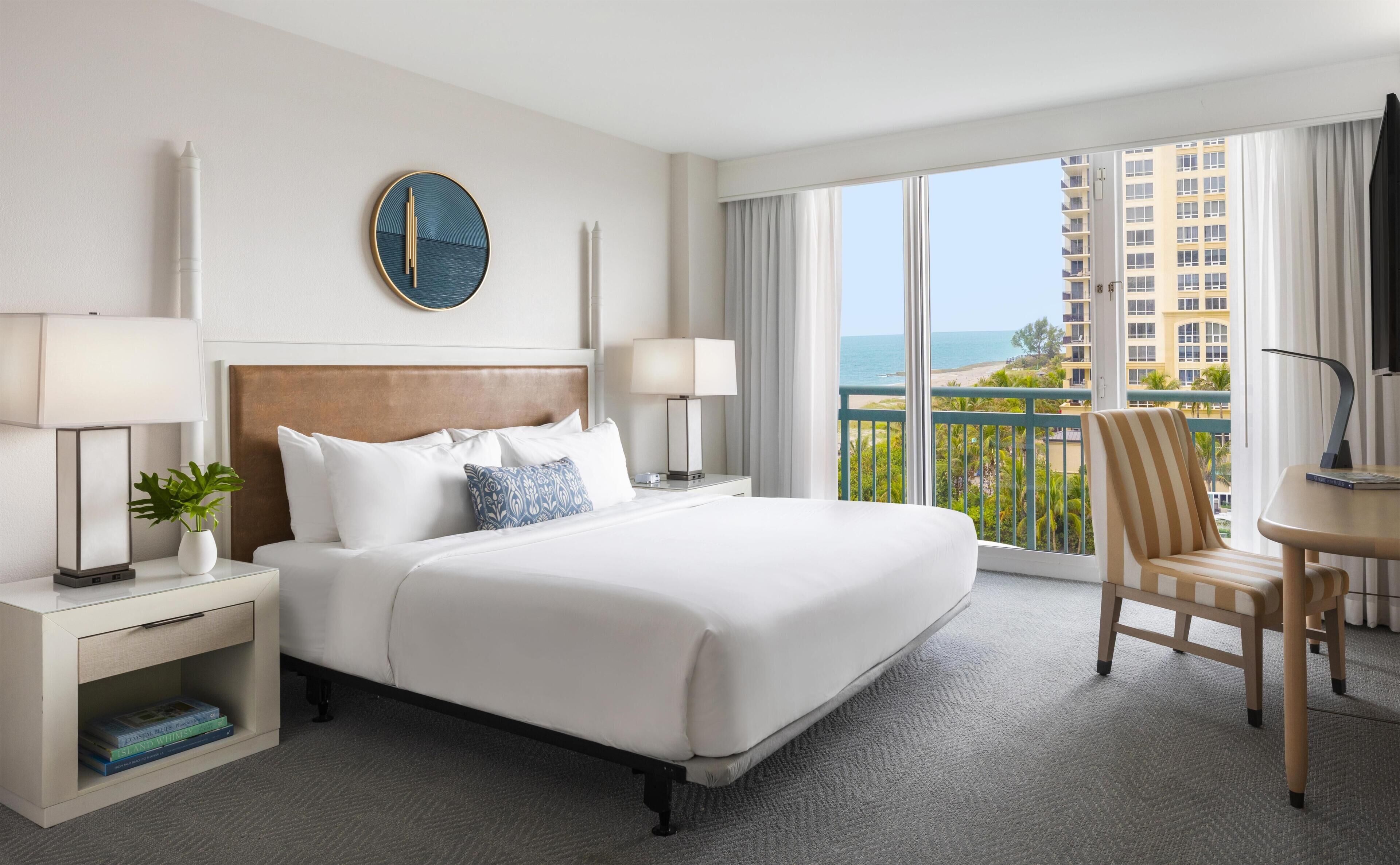 A hotel room with a large bed, two bedside tables with lamps, a view of the ocean, and a balcony with a chair.