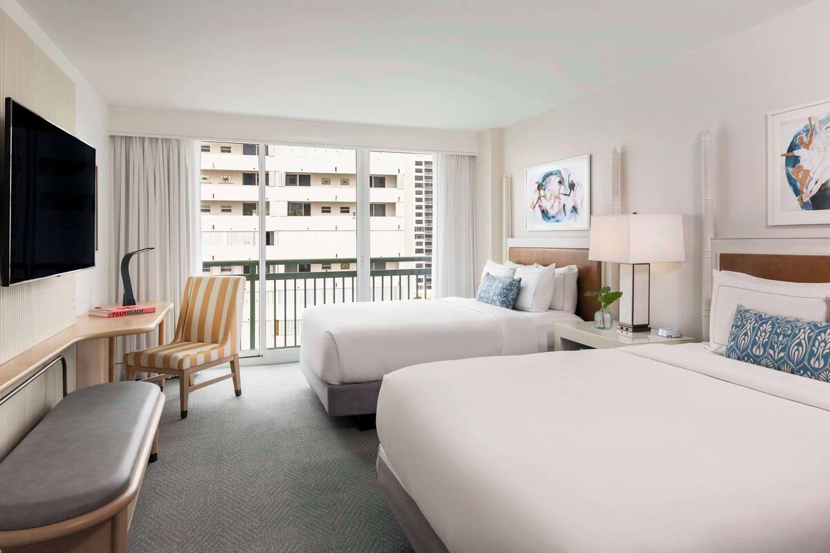 A hotel room with two beds, a desk, a chair, a TV, and a balcony with a view of the city.