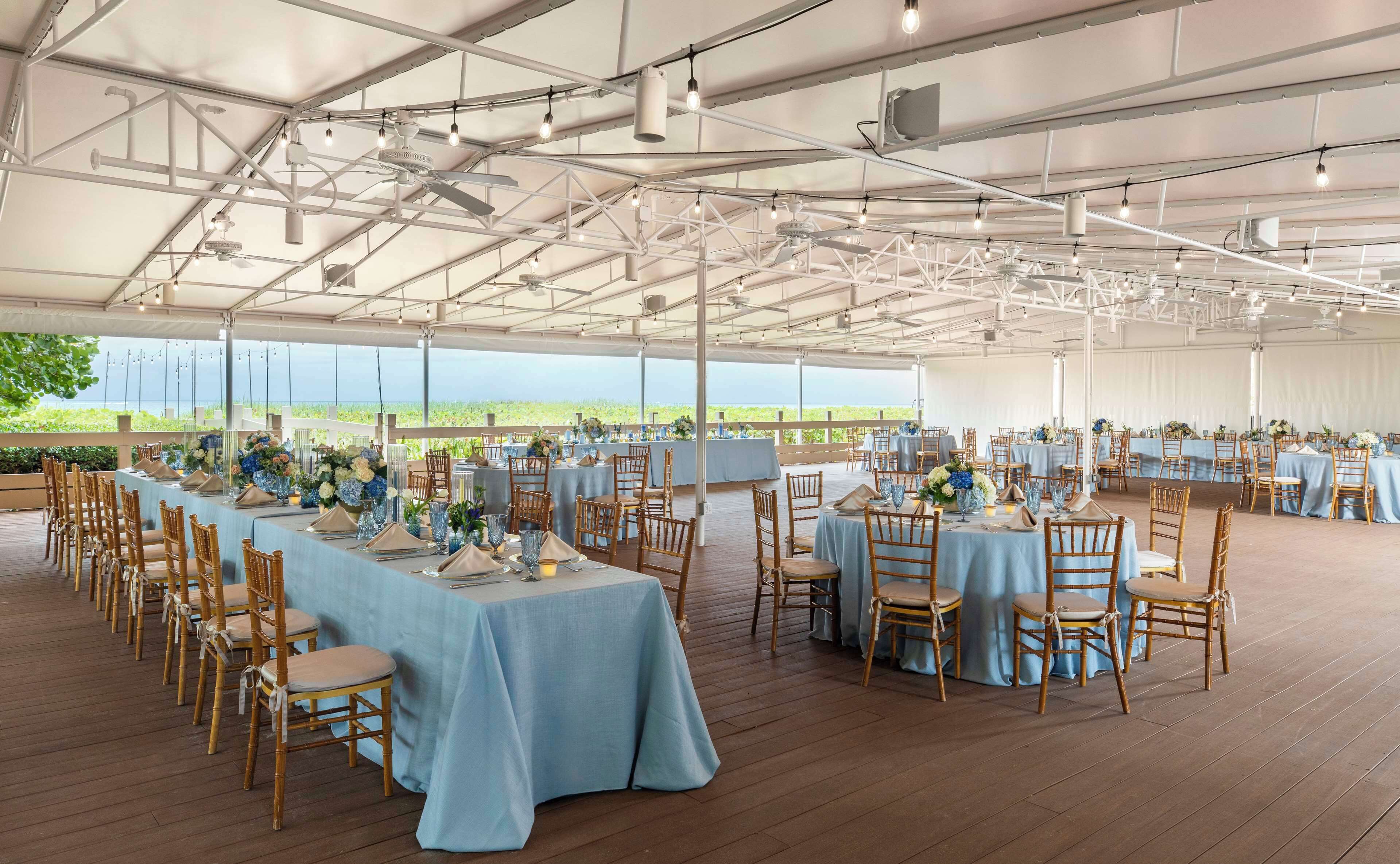 A large tented event space with tables set for a formal gathering, featuring blue tablecloths and wooden chairs.