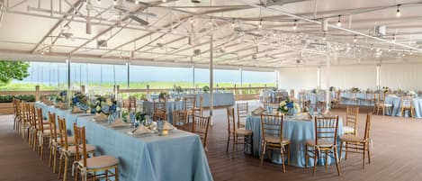 A large tented event space with tables set for a formal gathering, featuring blue tablecloths and wooden chairs.