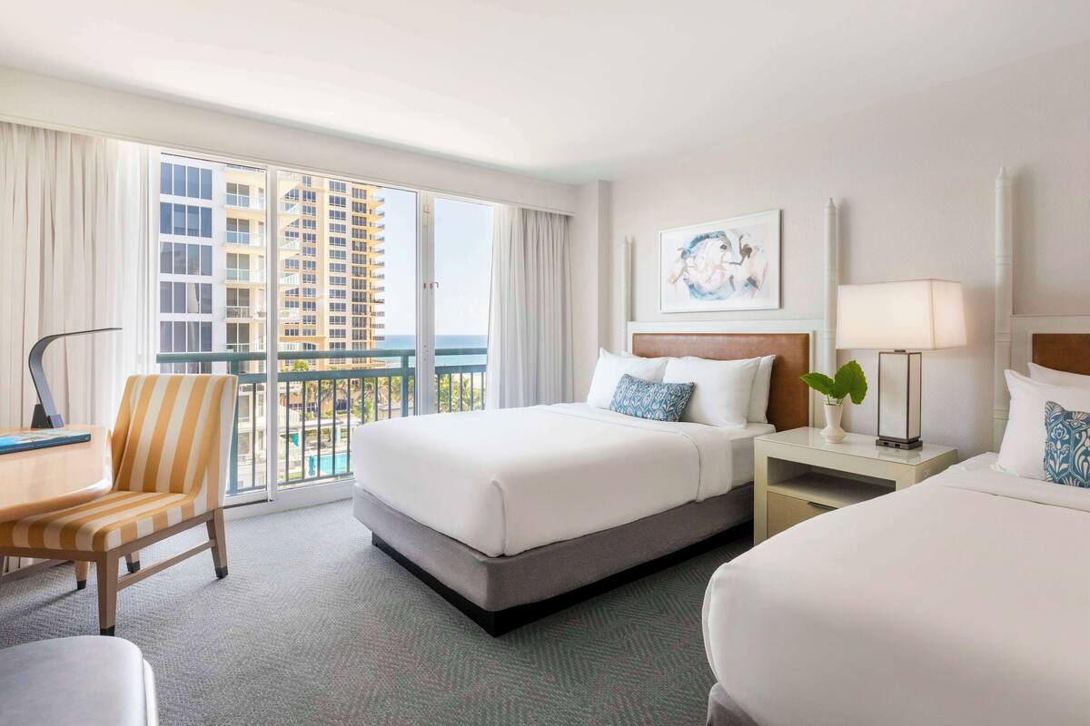 A hotel room with two beds, a chair, a desk, and a view of a high-rise building and the ocean.