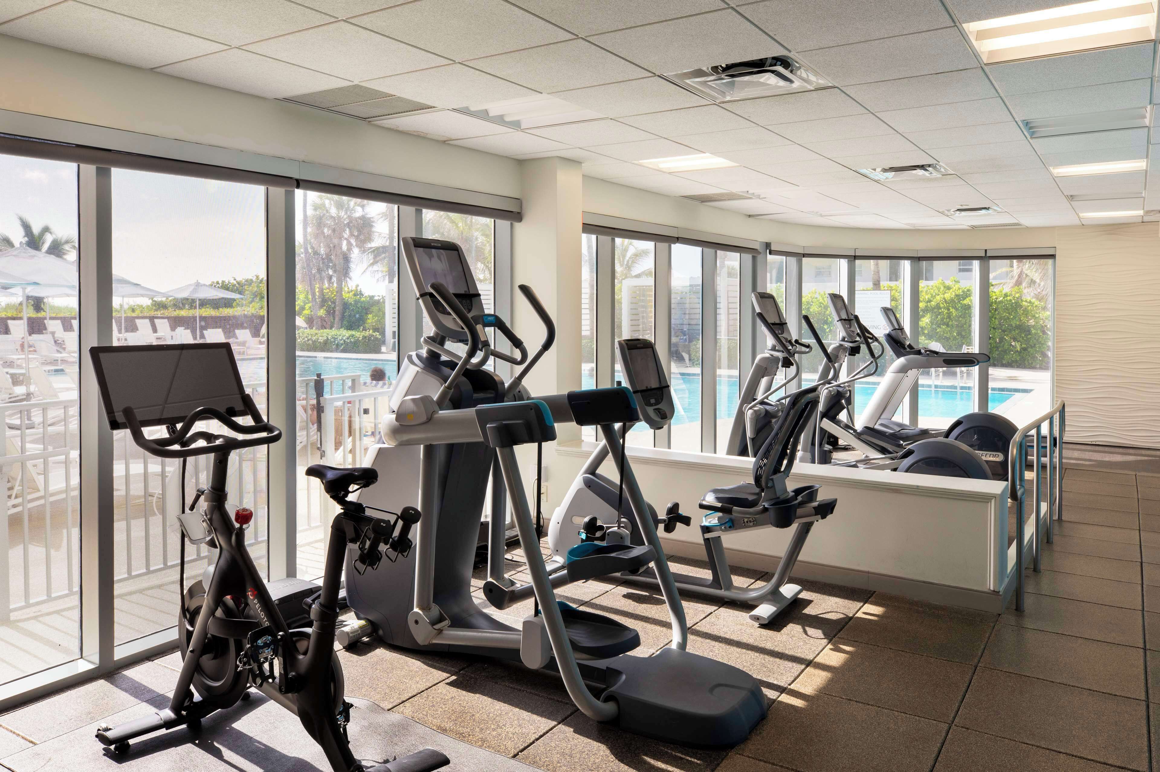 A gym with various exercise machines, large windows, and a view of a pool and palm trees.