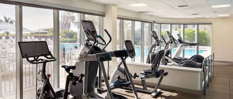 A gym with various exercise machines, large windows, and a view of a pool and palm trees.