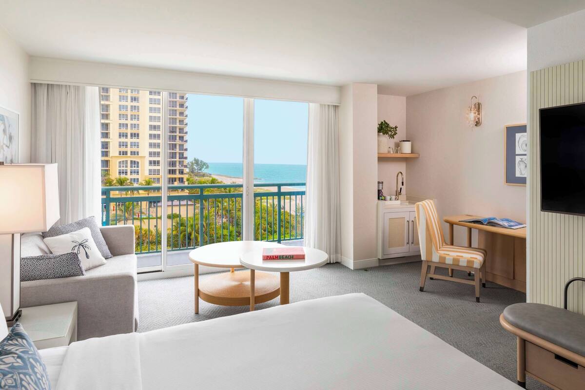 A hotel room with a large bed, a desk, a chair, a small table, a TV, a balcony with a view of the ocean and buildings.