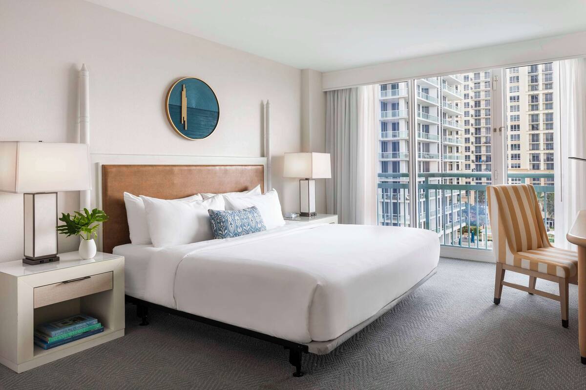 A hotel room with a large bed, two bedside tables, a chair, a view of a high-rise building, and a modern clock on the wall.