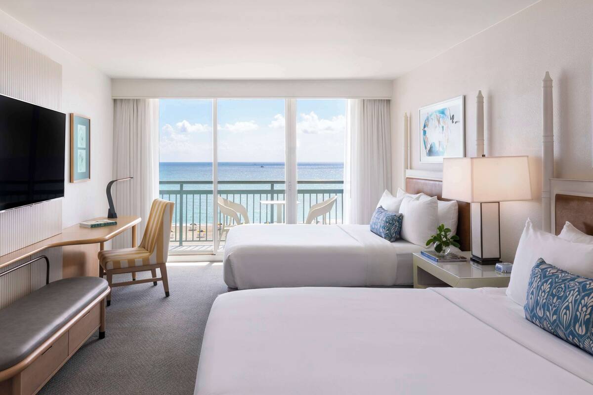 A hotel room with two beds, a desk, a TV, a balcony with a view of the sea, and a chair.