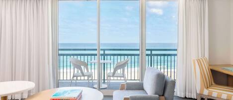 A hotel room with a large sliding glass door leading to a balcony with a view of the ocean. There are two chairs and a small table with a book on it.