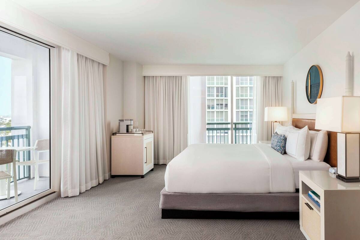 A hotel room with a large bed, a bedside table, a nightstand, a window with curtains, and a view of the city.