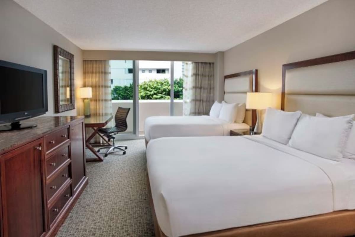 Quarto de hotel com duas camas, uma escrivaninha, televisão e vista para o verde lá fora.