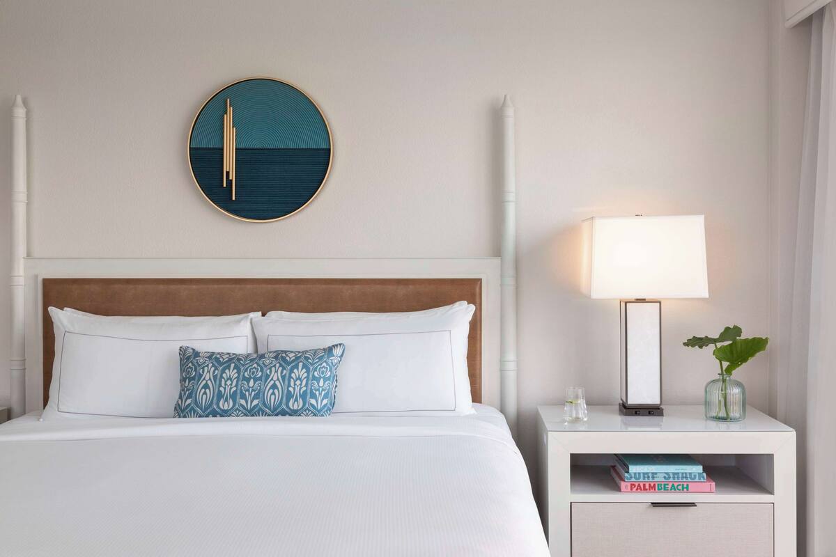 A neatly made bed with white linens and a patterned pillow, a round wall decoration, a bedside lamp, and a nightstand with books and a small plant.