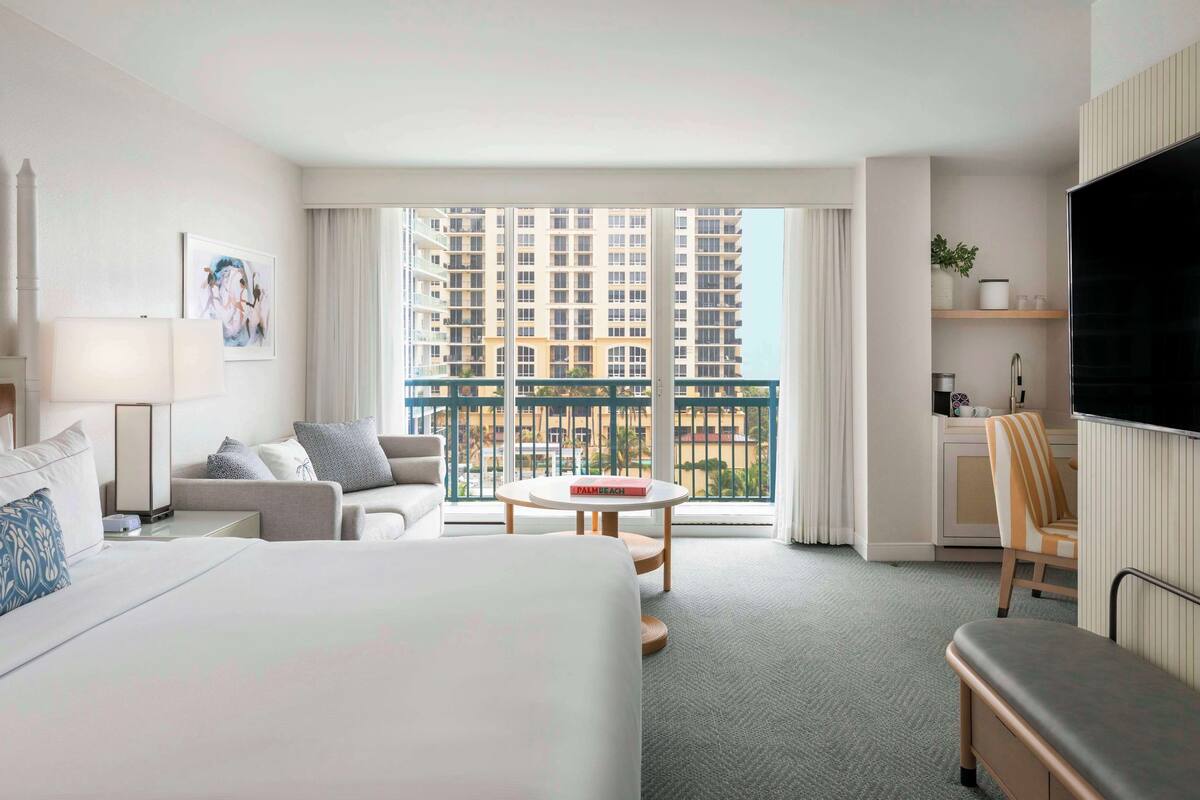 A hotel room with a bed, a sofa, a small table, a TV, a balcony with a view of high-rise buildings, and a desk with a chair.