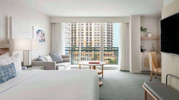 A hotel room with a bed, a sofa, a small table, a TV, a balcony with a view of high-rise buildings, and a desk with a chair.