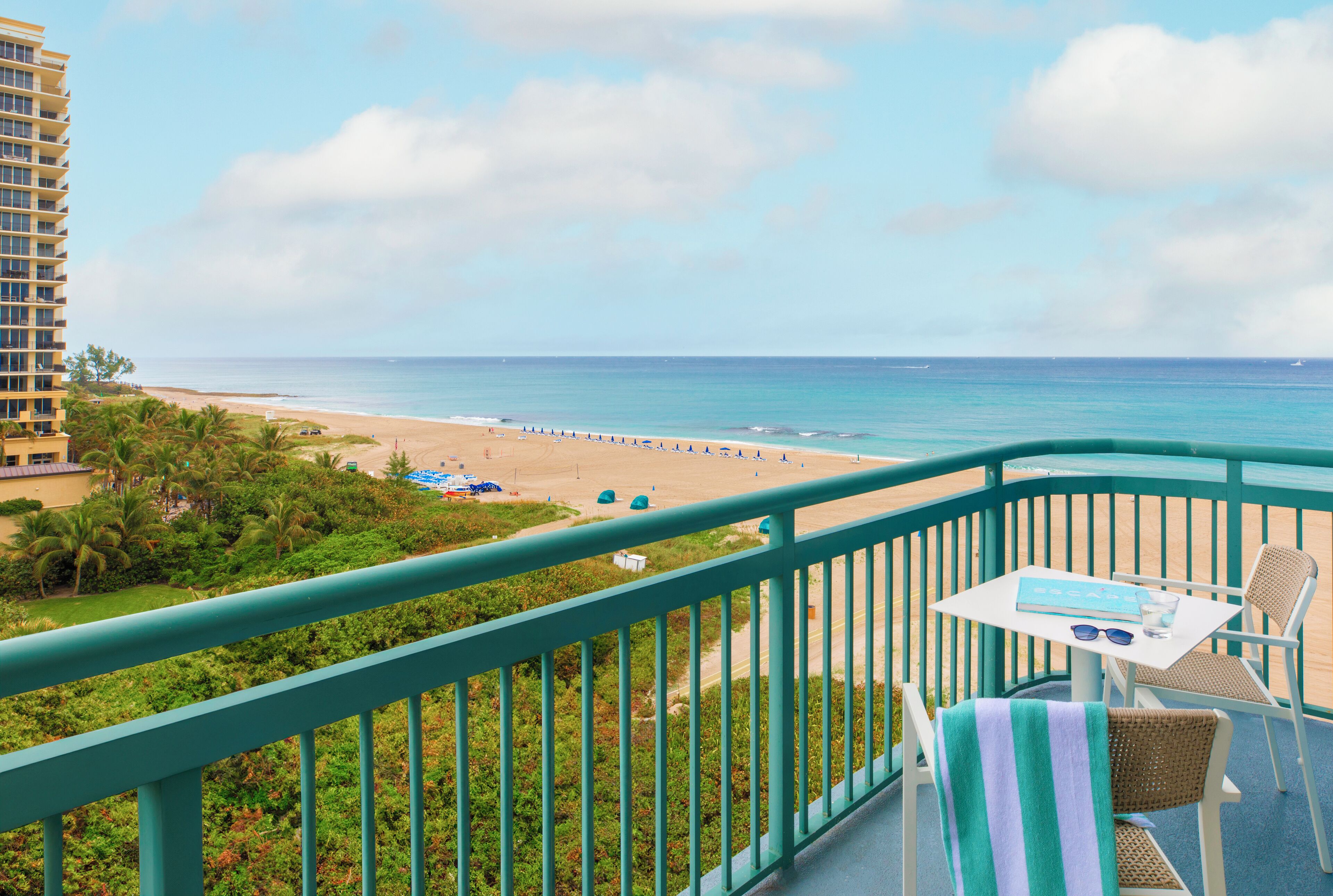 A balcony with a view of a beach and ocean. There is a table with a book and sunglasses on it.