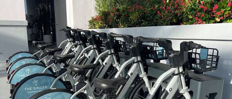 A row of stationary bikes with 'The Singer' branding, parked against a building facade with a white wall and greenery.