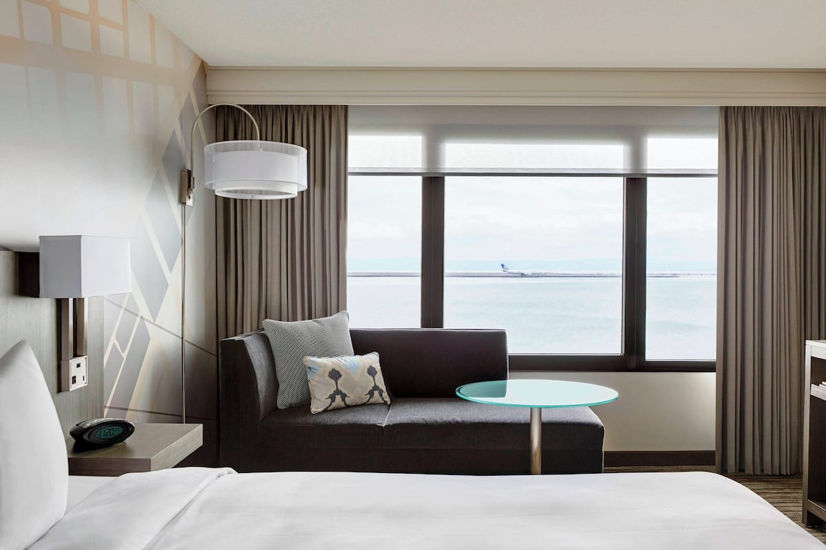 A hotel room with a bed, a sofa, a small round table, a lamp, and a view of the sea through large windows.