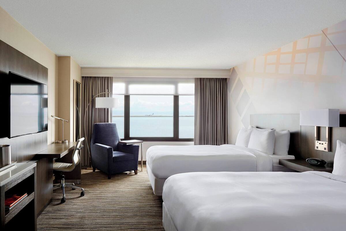 A hotel room with two beds, a desk with a chair, a TV, and a view of the sea.