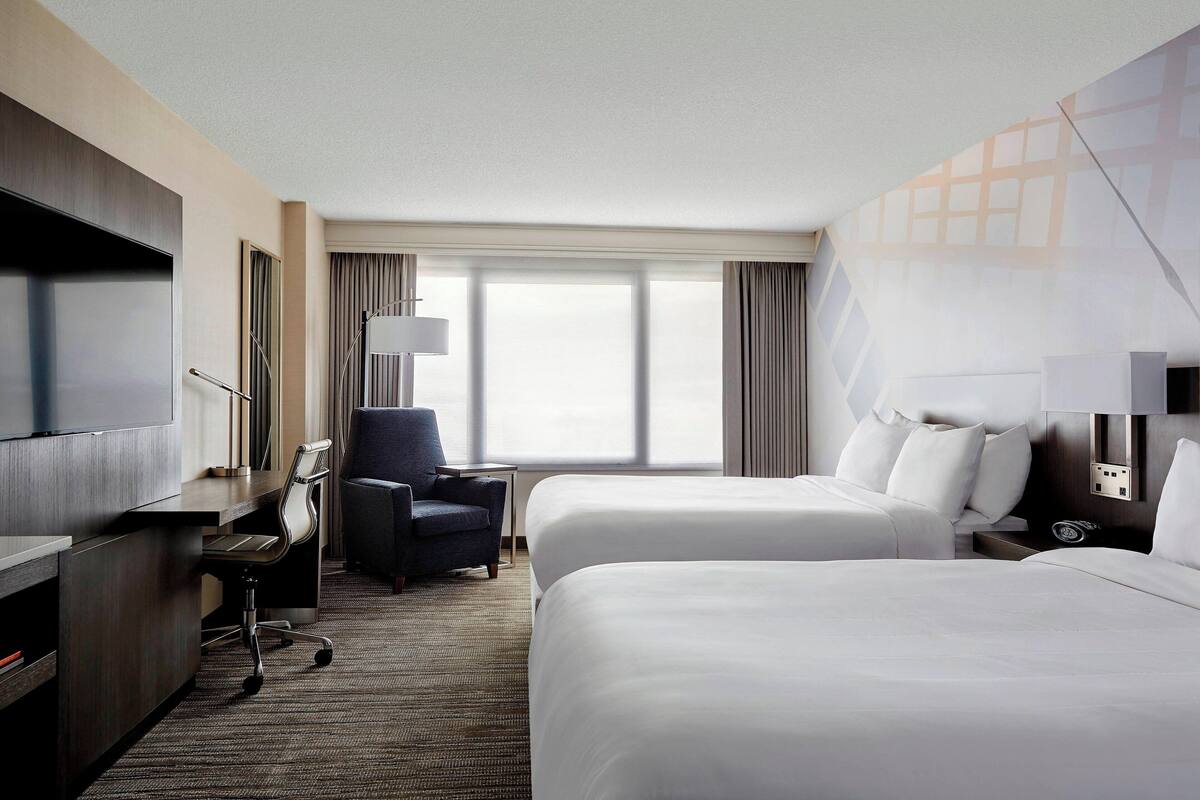 A hotel room with two beds, a desk, a chair, a TV, and a large window with curtains.