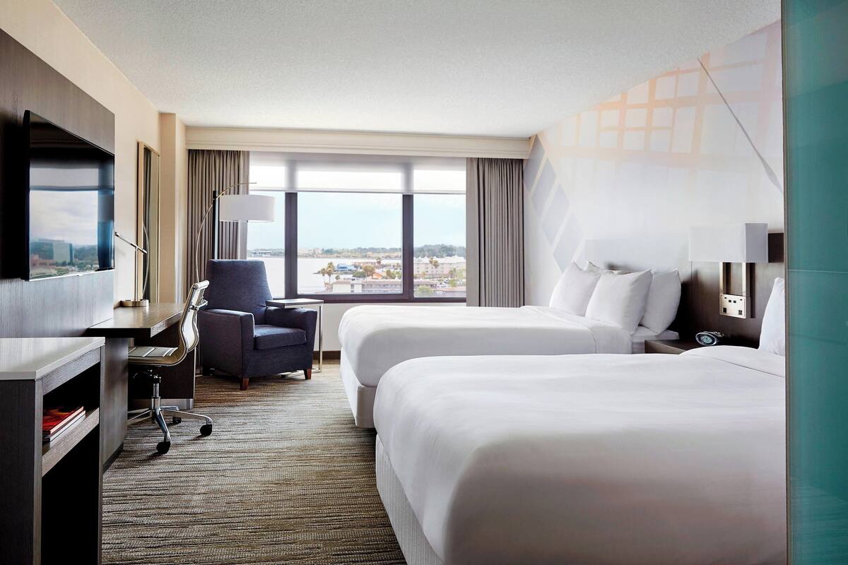 A hotel room with two beds, a desk, a chair, a TV, and a large window offering a city view.