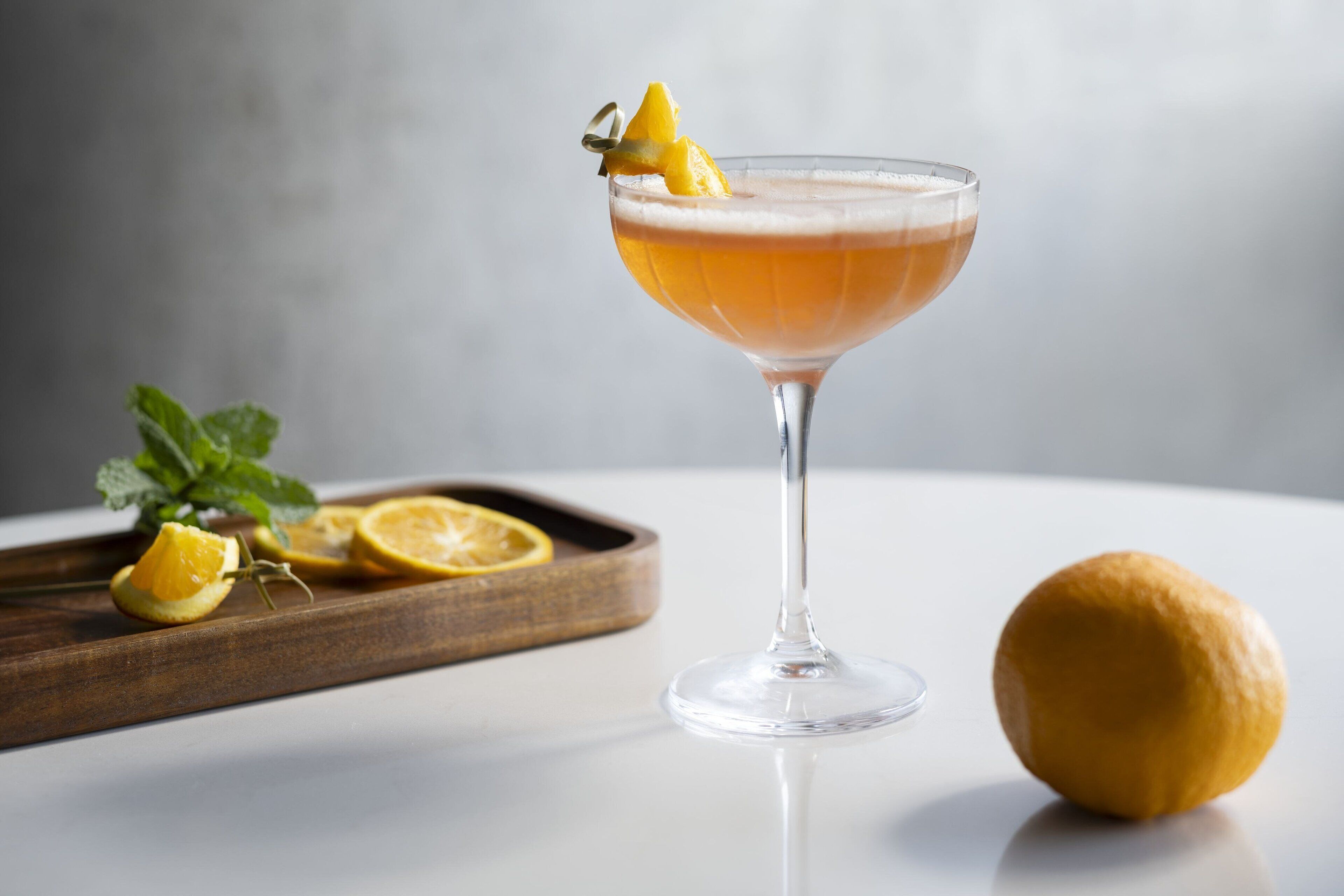 A cocktail in a glass with a lemon garnish, a wooden tray with lemon slices, and a whole lemon on a white surface.