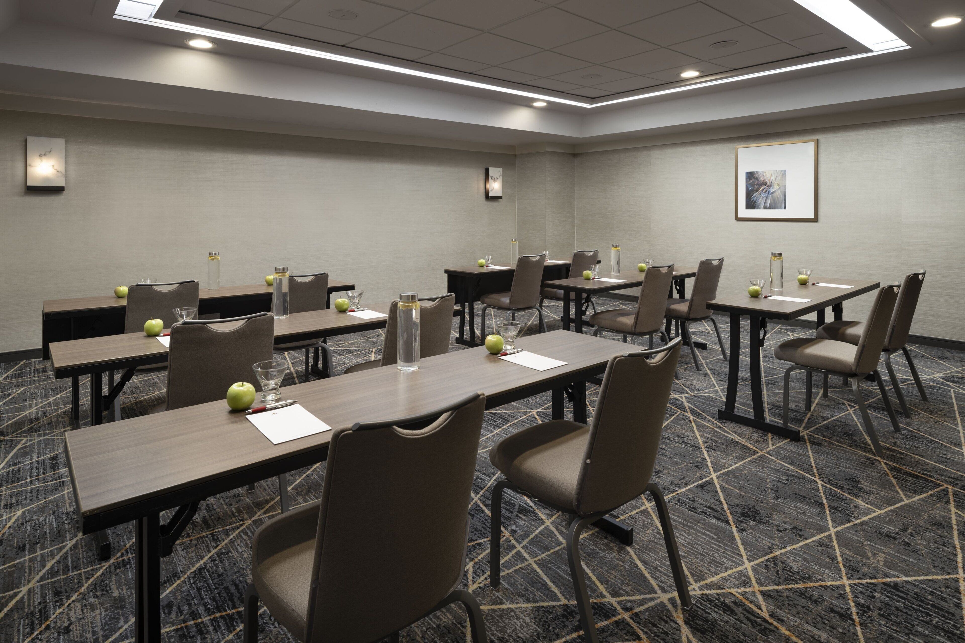 Un salón de conferencias con varias mesas y sillas, equipado con botellas de agua y manzanas, y un cuadro en la pared.