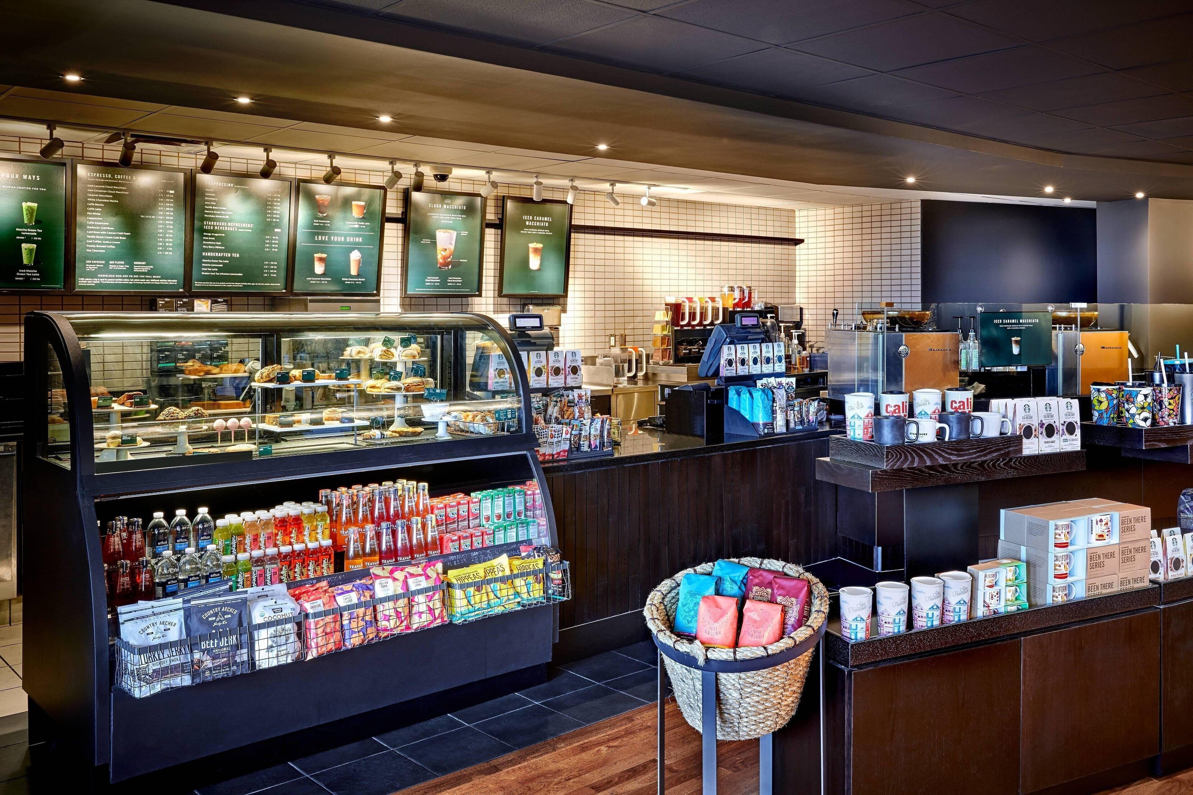 A coffee shop interior with a display case of pastries, a beverage cooler, and a counter with coffee machines and packaged goods.